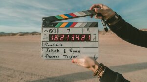 eine von zwei Händen geöffnete Filmklappe vor natürlichem Hintergrund: Sandboden, Sträucher, Himmel