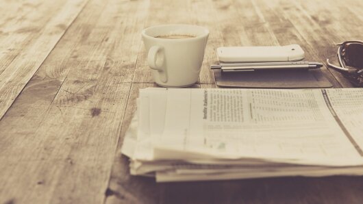 Zeitung, Kaffeetasse und Smartphone liegen auf einem Holztisch.