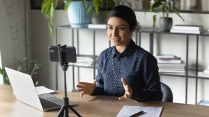 Eine junge Frau im digitalen Gespräch vor ihrem Smartphone, Laptop und Papier mit Stift liegen neben ihr.
